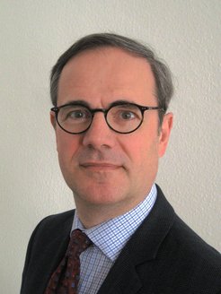 Grauer Anzug mit einem karierten Hemd und einer gemusterten Krawatte, vor einer hellen Wand.