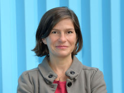 Kurzhaarige Frau mit brauner Jacke, rotem Oberteil und kleinem Anhänger, vor hellblauem, geripptem Hintergrund.