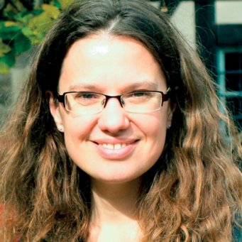 Dr. Olga Malets, Professur für Environmental Governance, Albert-Ludwigs-Universität Freiburg Braunes, lockiges Haar fällt über die Schultern eines Individuums vermutlich weiblichen Geschlechts. Es trägt eine unauffällige Brille und lächelt freundlich dem Betrachter entgegen, deutet auf ein friedvolles Gemüt hin. Im Hintergrund sind grüne Blätter und Teile eines Gebäudes zu sehen.