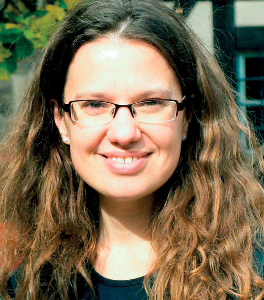 Braunes, lockiges Haar fällt über die Schultern eines Individuums vermutlich weiblichen Geschlechts. Es trägt eine unauffällige Brille und lächelt freundlich dem Betrachter entgegen, deutet auf ein friedvolles Gemüt hin. Im Hintergrund sind grüne Blätter und Teile eines Gebäudes zu sehen.