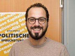 Ein gelbes Plakat im Hintergrund enthält Begriffe wie "Wahrheit", "Kapitalismus", "Globalisierung" und "Demokratie". Unten ist eine regelmäßige politische Sprechstunde angekündigt. Im Vordergrund ist ein freundlich lächelnder, schwarzhaariger, barttragender, brillenbewehrter junger Mann zu erkennen.