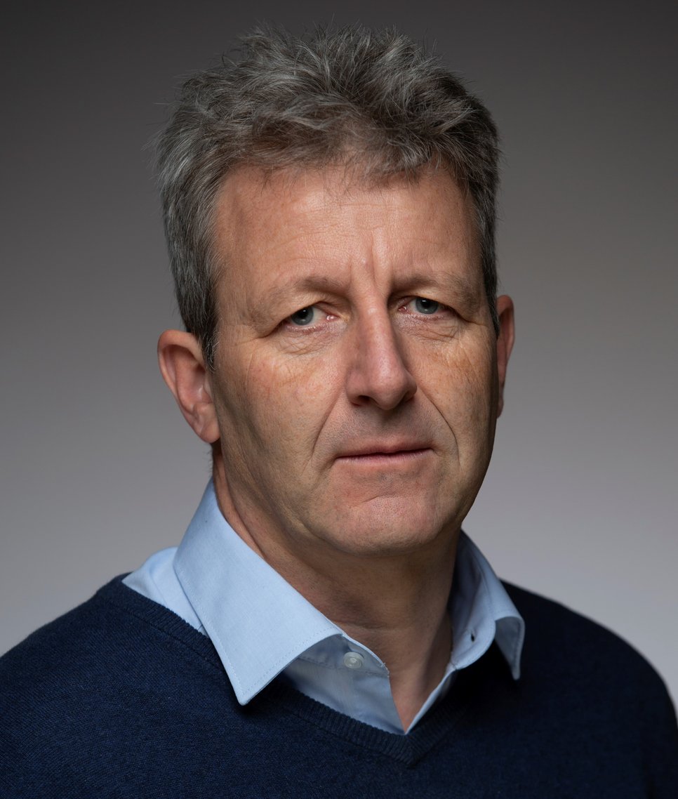 Individuum mit kurzem, grauem Haar, einem hellblauen Hemd mit offenem Kragen und dunklem Pullover ist vor neutralem Hintergrund zu erkennen. Individuum blickt konsterniert, möglicherweise übermüdet oder auch entgeistert. Der Mund ist geschlossen und deutet keinerlei Lächeln an.