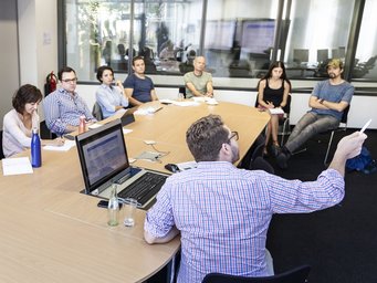 In a modern conference room, a group of eight people are discussing around an oval table, while one person is present with a laptop and notes.