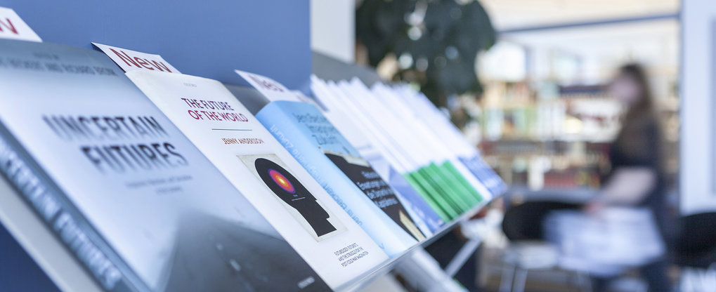 Eine Reihe von Büchern, die in einem Regal präsentiert werden. Die Titel umfassen "Uncertain Futures" und "The Future of the World". Im Hintergrund ist eine unscharfe Bibliotheksumgebung mit einer Person zu sehen.