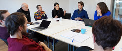 In a bright room, individuals are seated at a table, actively exchanging ideas, with laptops, notebooks, and coffee cups scattered around, indicating a collaborative work session.