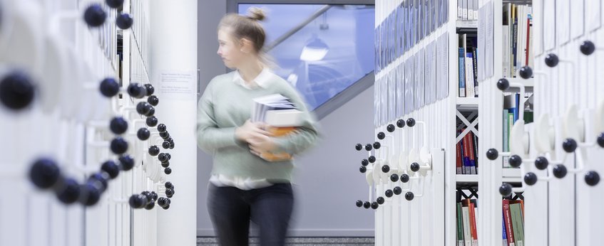 An individual moves through a library corridor, holding multiple books, surrounded by mobile shelving units with visible book spines.