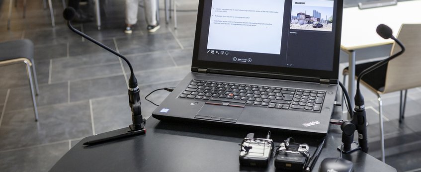 Conference setup featuring a Lenovo ThinkPad laptop on a desk with two microphones and wireless audio transmitters, surrounded by chairs on a tiled floor.