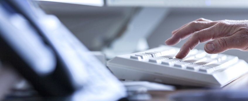A close-up image shows a person's hand actively typing on a white keyboard, while a phone is blurred in the foreground, suggesting an office environment.