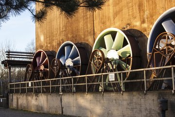Eine Reihe von vier großen Ventilatoren in verschiedenen Farben, montiert an einer Betonwand, umgeben von einem Geländer. Ein Baumast im Vordergrund.