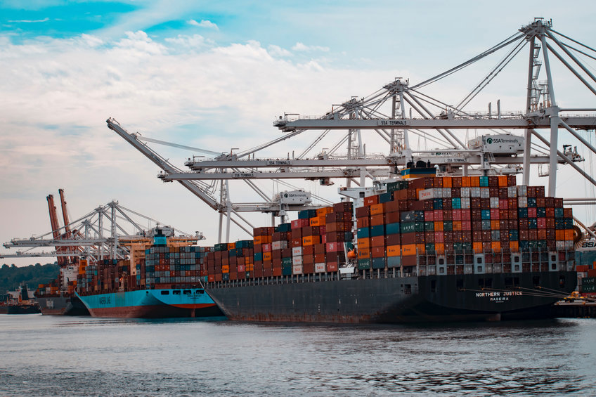 Kapitalismus braucht Wachstum – aber es gibt kein Patentrezept Im Vordergrund befinden sich zwei beladene Containerschiffe in einem Hafen. Mehrere Kräne erstrecken sich über die Schiffe, während im Hintergrund weitere Schiffe am Kai zu sehen sind.