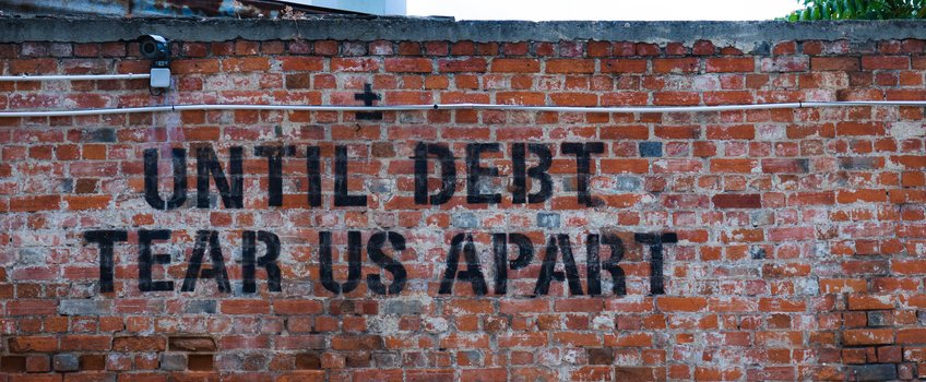 Die deutsche Bevölkerung lehnt Eurobonds nicht grundsätzlich ab Auf einer roten Backsteinmauer ist ein schwarzer Schriftzug zu sehen, der den Spruch „Until debt tear us apart“ enthält. In der oberen Ecke ist eine Überwachungskamera angebracht.