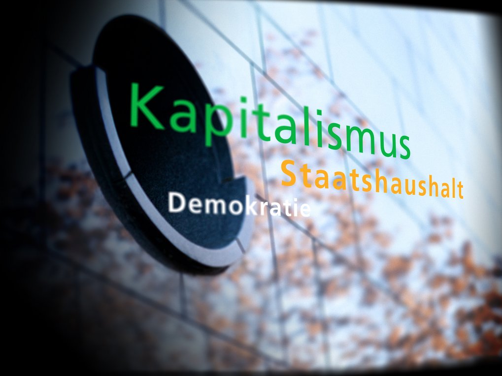 Glasfassade überzogen mit den farbigen Schriftzügen "Kapitalismus", "Staatshaushalt", "Demokratie"