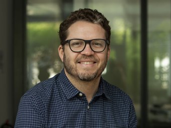 Ein vermutlich männliches, noch recht junges Exemplar der Gattung Homo sapiens intellectualis ist auf dem Foto zu erkennen – vor einem großen Glasfenster, wodurch verschwommenes Grün sickert, sitzend oder stehend. Es trägt ein blau-weiß kariertes Hemd mit offenem Kragen, auf der Nase eine Brille, die der verfeinerten optischen Wahrnehmung zweckdienlich ist. Das Individuum hat den Mund zu einem als optimistischem Lächeln interpretierbaren Gestus geöffnet. Nicht unerwähnt darf bleiben, dass das Individuum einen bereits ausgeprägten Bartwuchs aufweist, das auf Rasierverzicht von mindestens zweiwöchiger Dauer hindeutet.