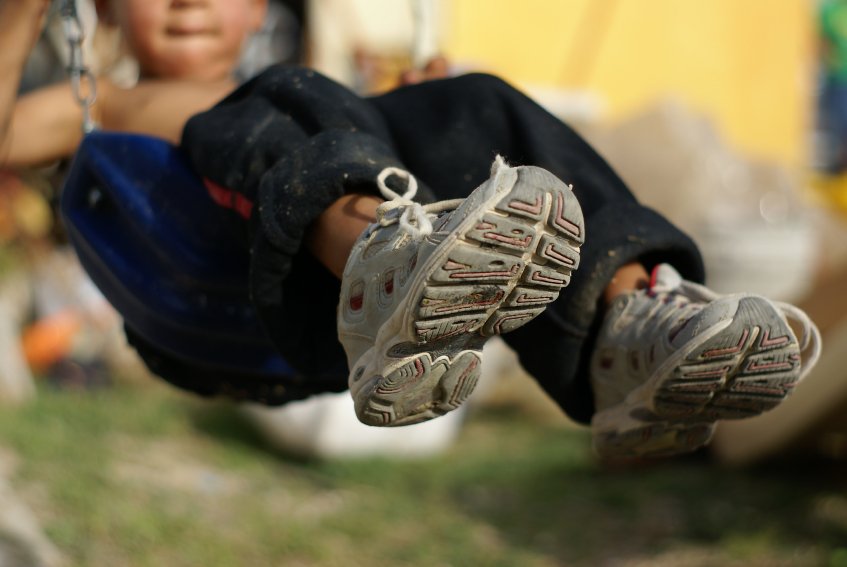 Children Are Not For Sale: Intercountry Adoption as a Field of Conflict: The Absence of Market Characteristics Close-up of a child's feet in white sneakers, sitting on a blue swing, with blurred outdoor background.