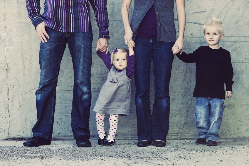 Doppelpaariges Menschenbündnis A group of four, comprising two adults and two children, stand side by side against a concrete backdrop. The adults, dressed in casual attire, hold the children's hands, conveying a sense of togetherness and familial bond.