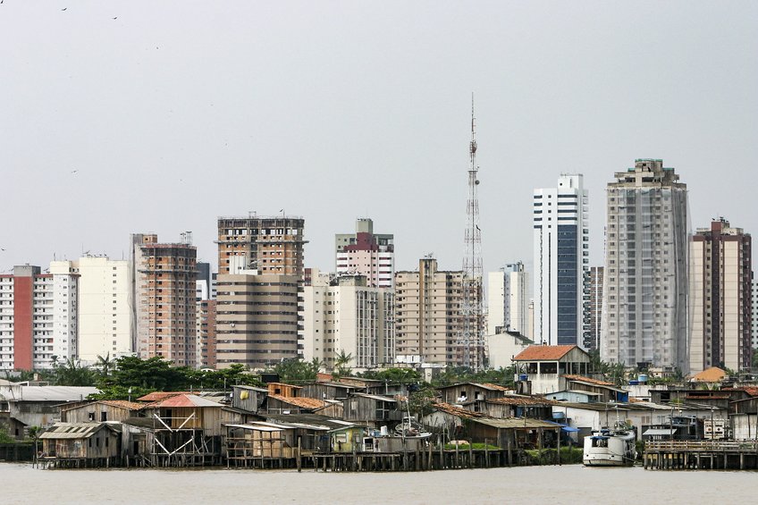 Stadtlandschaft mit modernen Hochhäusern und informellen Hütten Urban landscape featuring a mix of modern high-rise buildings and informal riverside shacks, with a prominent communication tower in the background.