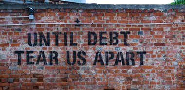 On a red brick wall, in large black letters, it reads "Until debt tear us apart". A surveillance camera is mounted at the top left, with cables running beside it.