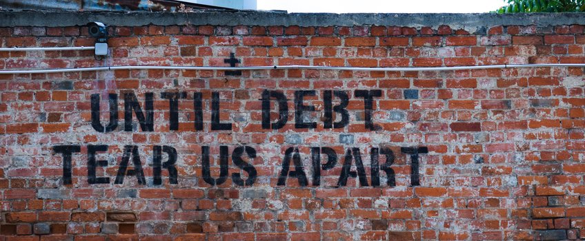 On a red brick wall, in large black letters, it reads "Until debt tear us apart" On a red brick wall, in large black letters, it reads "Until debt tear us apart". A surveillance camera is mounted at the top left, with cables running beside it.