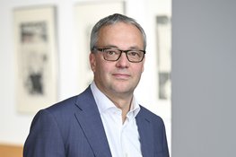 Person wearing a dark blue suit and white shirt, standing in front of a light background with blurred images on the wall.
