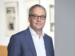 Person wearing a dark blue suit and white shirt, standing in front of a light background with blurred images on the wall.