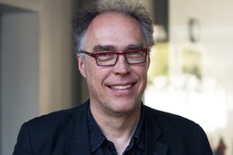 Person with curly gray hair, wearing a dark blue shirt and a dark jacket. Background shows blurred interior elements.