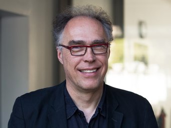 Person with curly gray hair, wearing a dark blue shirt and a dark jacket. Background shows blurred interior elements.