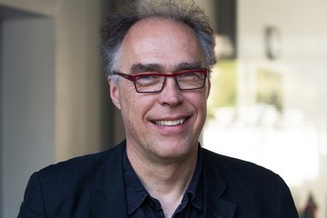 Person with curly gray hair, wearing a dark blue shirt and a dark jacket. Background shows blurred interior elements.