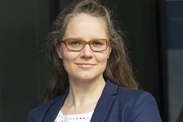 A person with long hair wears a dark blue blazer over a white, polka-dot top, in front of a dark background.