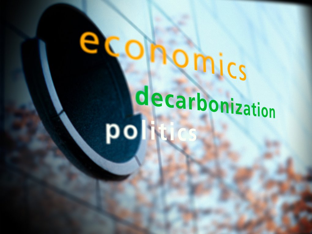 Text "economics" in orange, "decarbonization" in green, and "politics" in white on a blurred background with abstract elements.