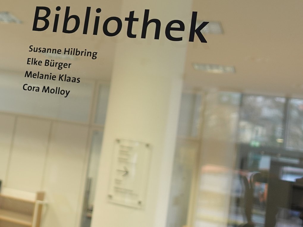 Glastür der Bibliothek zeigt die Namen Susanne Hilbring, Elke Bürger, Melanie Klaas, Cora Molloy mit verschwommenem Blick ins Innere.
