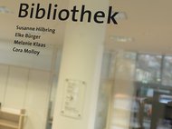 Glass door of library shows names Susanne Hilbring, Elke Bürger, Melanie Klaas, Cora Molloy with blurred interior view.