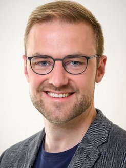 Ein Individuum mit grauem Jackett mit Fischgrätenmuster ist auf dem Foto zu erkennen. Es hat hellbraunes, kurzes Haar, trägt eine Brille zwecks besserer optischen Erfassung der Umgebung. Das Individuum lächelt freundlich den Betrachtenden an, der Mund ist halb geöffnet, ein Bartanflug deutet auf eine mindestens viertägige rasurfreie Periode hin.