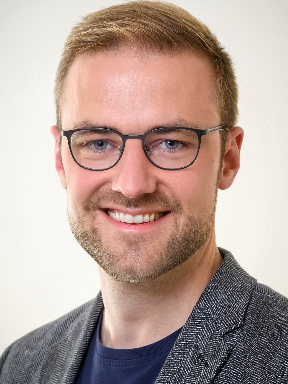 Ein Individuum mit grauem Jackett mit Fischgrätenmuster ist auf dem Foto zu erkennen. Es hat hellbraunes, kurzes Haar, trägt eine Brille zwecks besserer optischen Erfassung der Umgebung. Das Individuum lächelt freundlich den Betrachtenden an, der Mund ist halb geöffnet, ein Bartanflug deutet auf eine mindestens viertägige rasurfreie Periode hin.