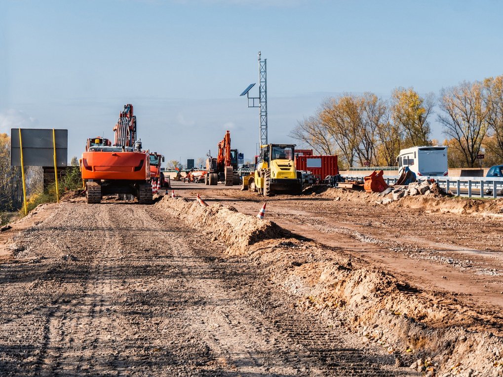 Eine Baustelle mit Baggern und Baugeräten auf einer unbefestigten Straße. Im Hintergrund sind Bäume und ein Wohnmobil zu sehen, während Bauarbeiten stattfinden. Die Sonne scheint und der Himmel ist klar.
