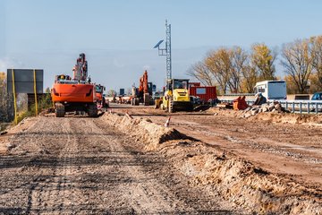 Eine Baustelle mit Baggern und Baugeräten auf einer unbefestigten Straße. Im Hintergrund sind Bäume und ein Wohnmobil zu sehen, während Bauarbeiten stattfinden. Die Sonne scheint und der Himmel ist klar.
