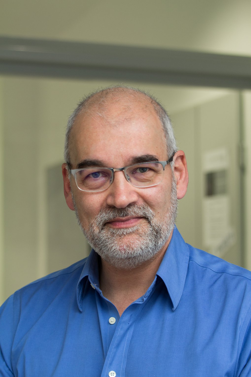 Eine Person mit blauem Hemd steht vor einer grauen Wand, im Hintergrund sind unscharfe Schilder an der Wand angebracht.