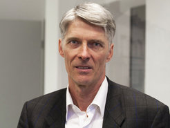 The photo shows an individual of the species Homo sapiens. He has gray hair, is wearing a dark suit and a white shirt with a collar that is open at the neck. The hair is already gray, but still full and shiny. The hair is parted on the right side and neatly combed. The mouth is half open, and the face shows the hint of a tentative smile that can be discerned with sufficient goodwill. The evidence suggests that the scene is set in an office.