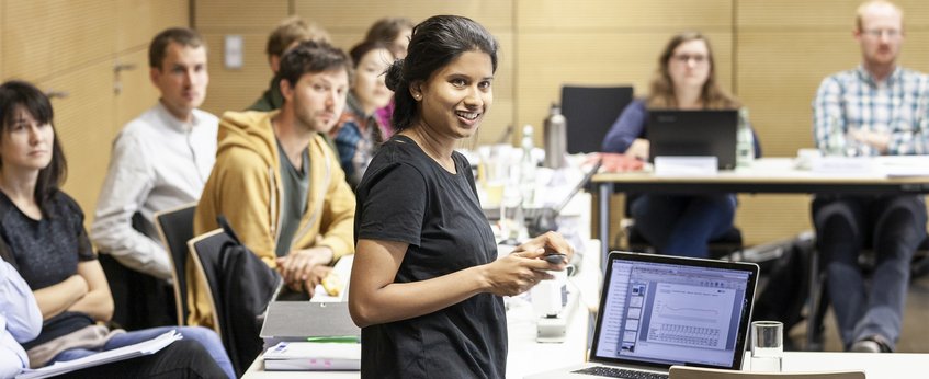 A speaker addresses a seated audience in a conference setting, with a laptop projecting graphs and charts for discussion.