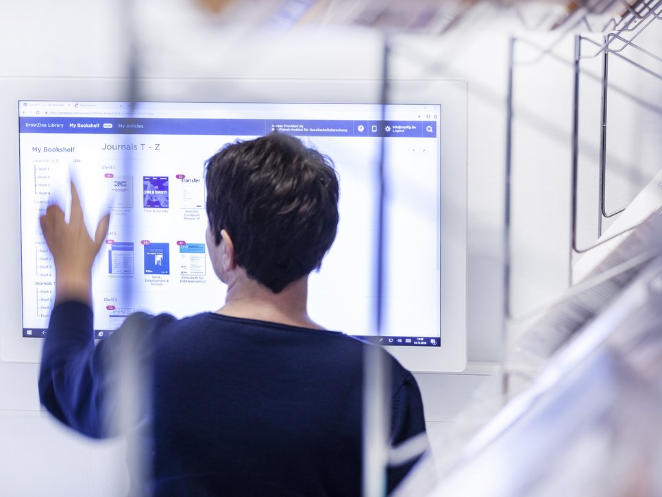 A person stands in front of a large touchscreen, navigating a digital library interface. Visible are journal covers and article listings, with shelves in the background.