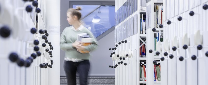 A blurred figure of a woman carrying books walks between white bookshelves in a library.