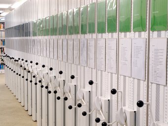 Eine Reihe mobiler Bibliotheksregale, die jeweils mit Kurbelgriffen zur manuellen Verstellung ausgestattet sind. Darüber sind Dokumente und grüne Ordner ausgestellt, die Kataloginformationen enthalten.
