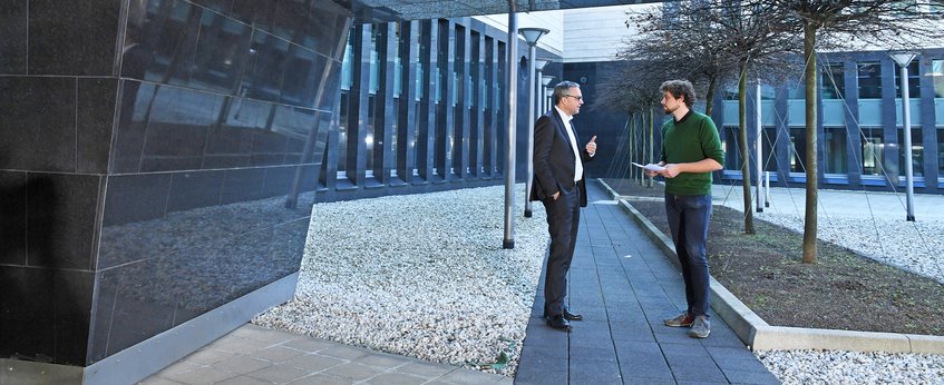 Two men are conversing outdoors in front of a modern building complex with sidewalks and trees.