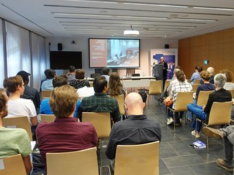 Mehrere Personen sitzen in Reihen und verfolgen aufmerksam die Rede eines Vortragenden, der auf einem Podium in einem modernen Konferenzraum steht. Auf einer großen Leinwand werden Präsentationsfolien zum Thema Arbeit gezeigt.