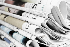 A stack of folded newspapers displays various headlines and images in a mixture of color and black and white, revealing details of headlines and articles.