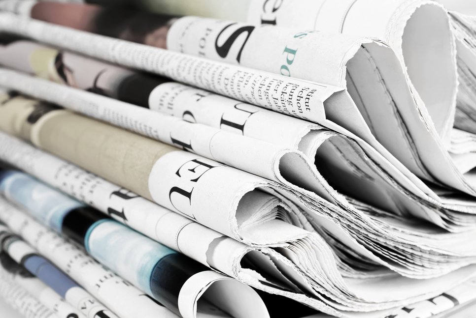 Ein Stapel gefalteter Zeitungen zeigt verschiedene Überschriften und Bilder in einer Mischung aus Farbe und Schwarzweiß, die Details von Schlagzeilen und Artikeln erkennen lassen.