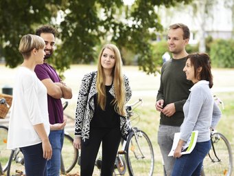Eine gemischte Gruppe von fünf Personen steht in einem Park und unterhält sich. Im Hintergrund sind zwei Fahrräder zu sehen, was auf ein zwangloses Treffen oder eine Pause während einer Radtour hindeutet. Die Umgebung ist entspannt, Bäume spenden Schatten.