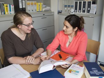 Two women are sitting at a table in an office, deep in discussion. Open books and various file folders are spread out in front of them. In the background, a shelf with more folders and books can be seen.