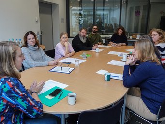 The administration team at one of the regular service group meetings. A meeting in a conference room with participants seated around a table, papers, notebooks, mugs, and bottles are present, indicating an organized discussion setting.