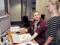 An office with two people, a desk full of documents, a computer with an open browser, and various office supplies.