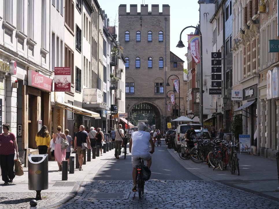 Cyclists ride and pedestrians walk along the busy shopping street, flanked by shops and a historic archway in the background.
